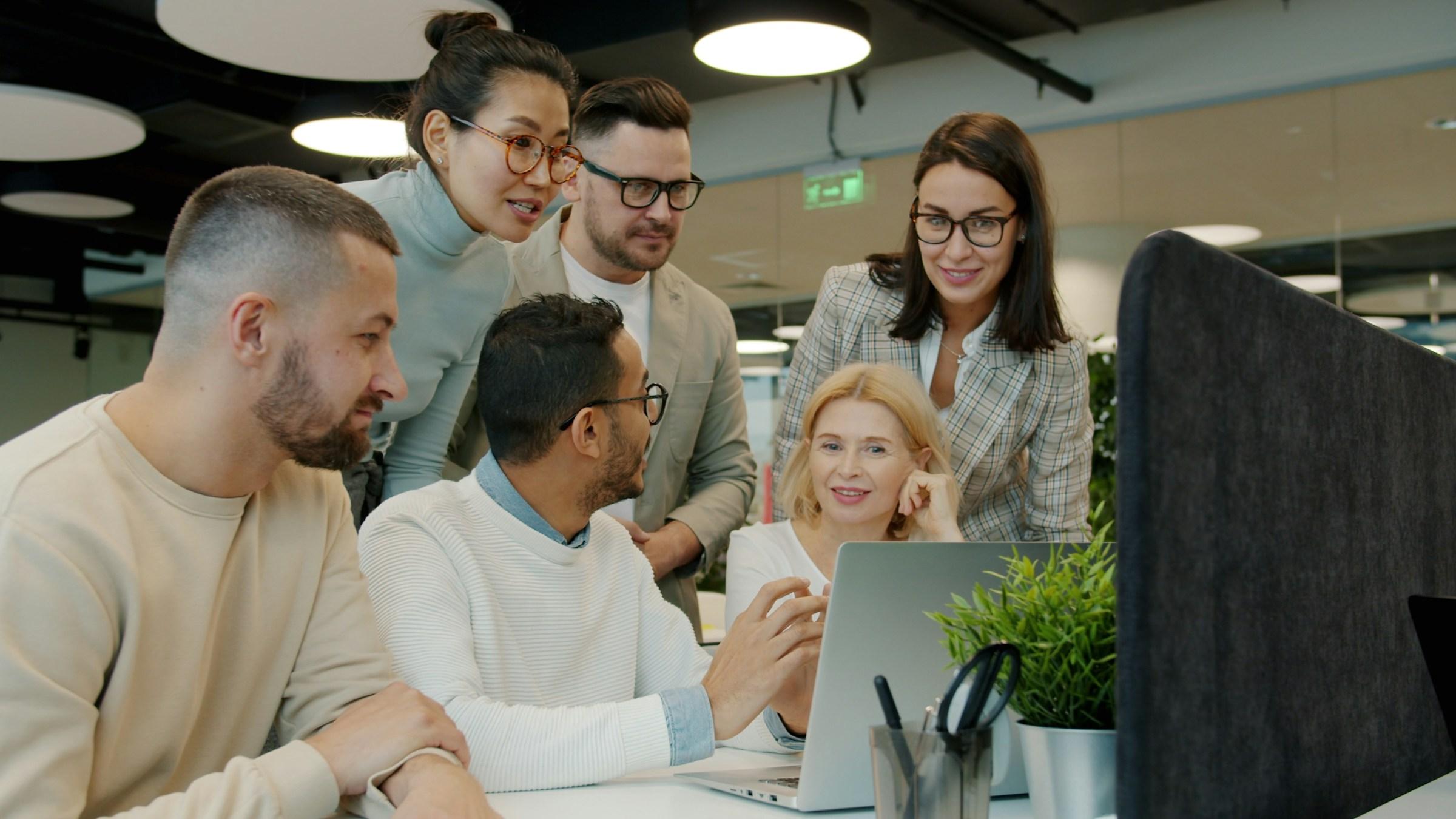 Six colleagues gathered around a laptop in a bright, modern office